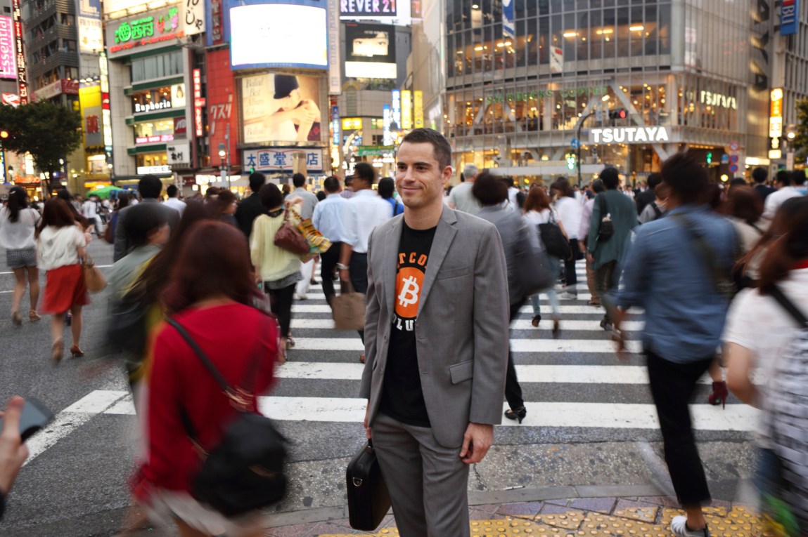 A man is wearing a suit jacket with a T-shirt underneath featuring the bitcoin logo. He is standing in the middle of a busy cosmopolitan intersection, and the many people walking around him are blurred.