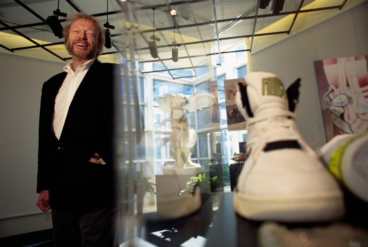A bearded man wearing a black suit and white shirt smiles at the camera. He is standing in a room filled with various art about shoes. A replica of the Niké of Samothrace statue is visible between him and a pair of white shoes.