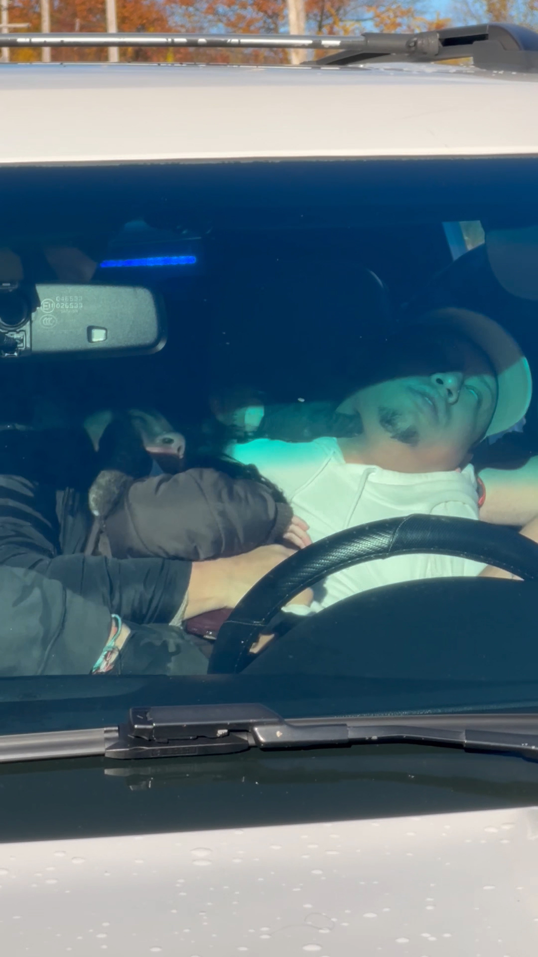 A man wearing a white shirt and baseball hat convulses in the driver’s seat of a car while a black-gloved hand presses into his neck.