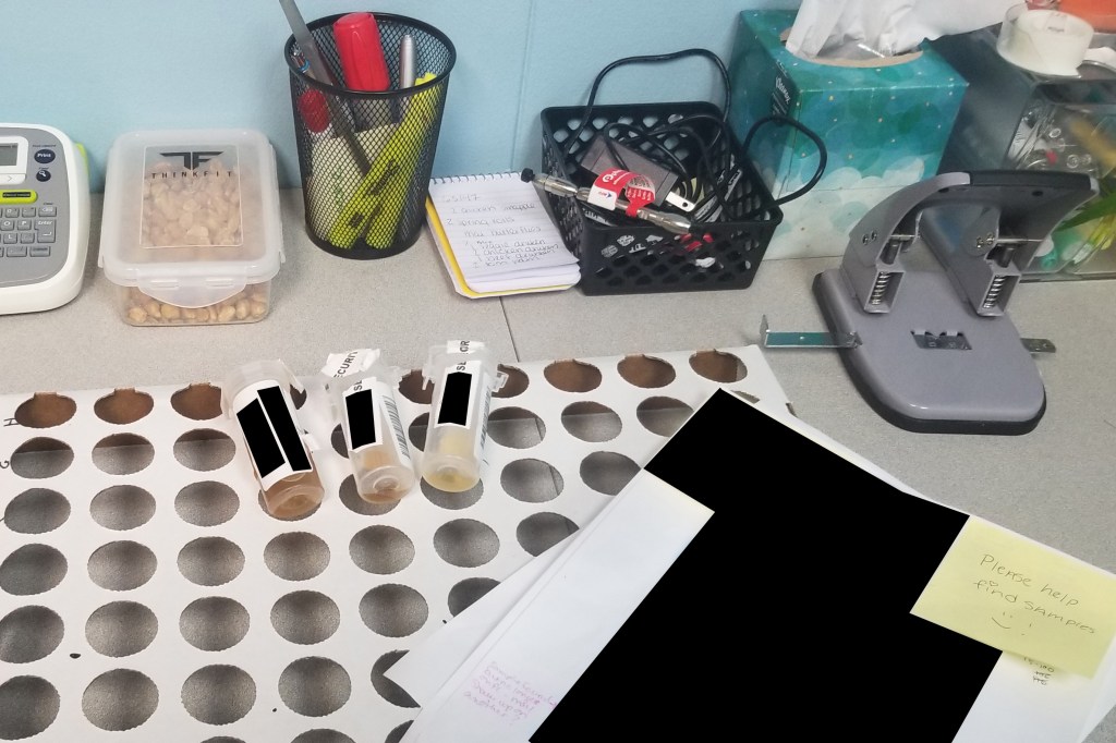 Three lab testing vials on a desk, alongside redacted documents, a container of nuts and office equipment.