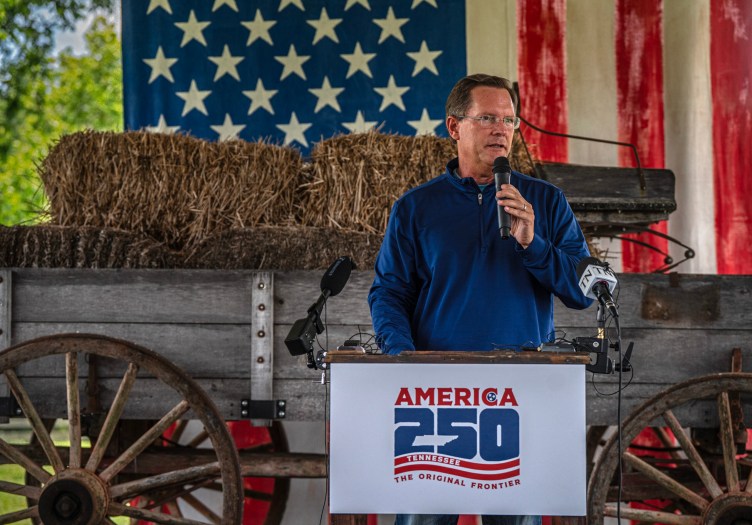 A man stands at a podium and speaks into a microphone in front of a wagon full of hay and a large American flag.