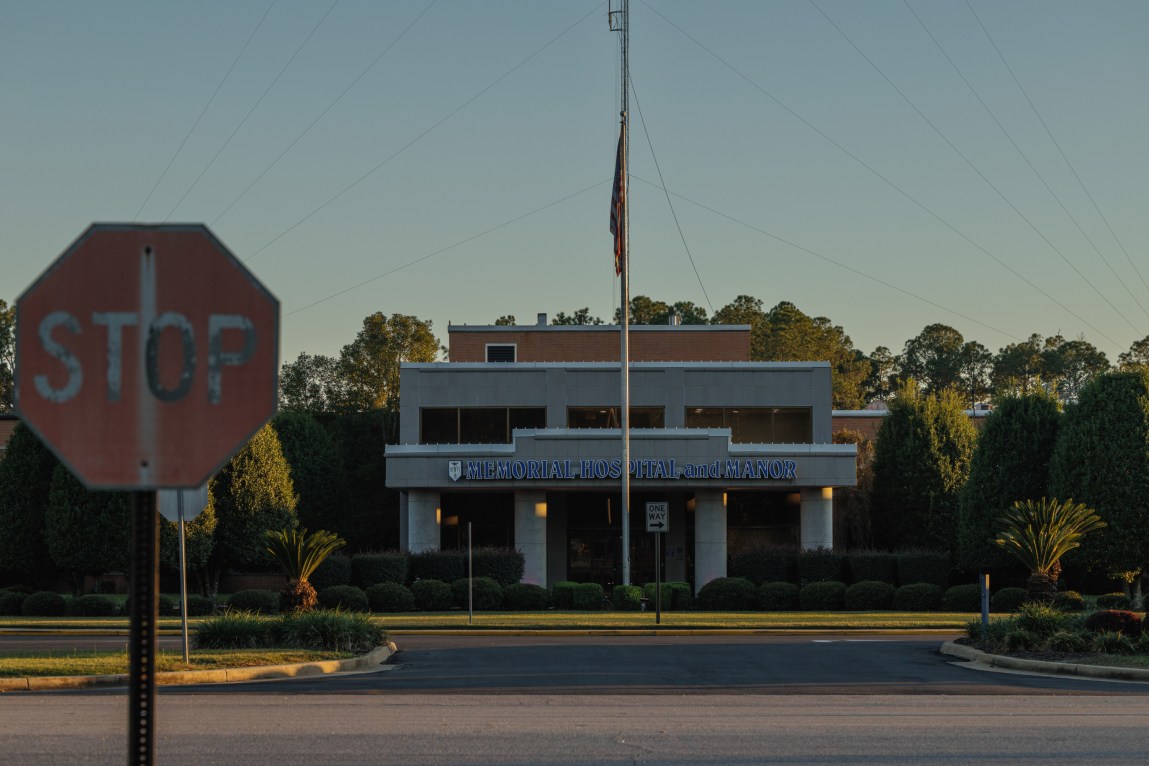 A faded stop sign stands in front of a building with the sign “Memorial Hospital and Manor.”