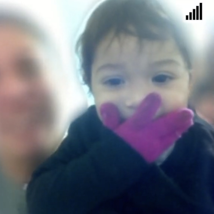 A toddler wears a jacket and a purple glove on her right hand, which covers her mouth. Behind her is a blurry woman smiling.