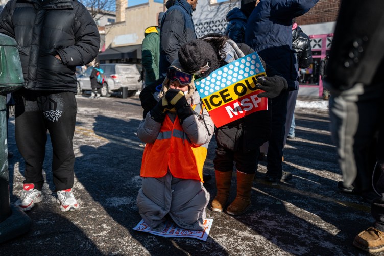 Des gens en vêtements d’hiver se tiennent dans une rue. Une femme portant un gilet orange et des lunettes de sécurité s'agenouille sur le sol, les mains jointes devant son visage. Une autre femme se penche pour passer ses bras autour de la femme agenouillée. Elle tient une pancarte indiquant « Ice Out MSP ».