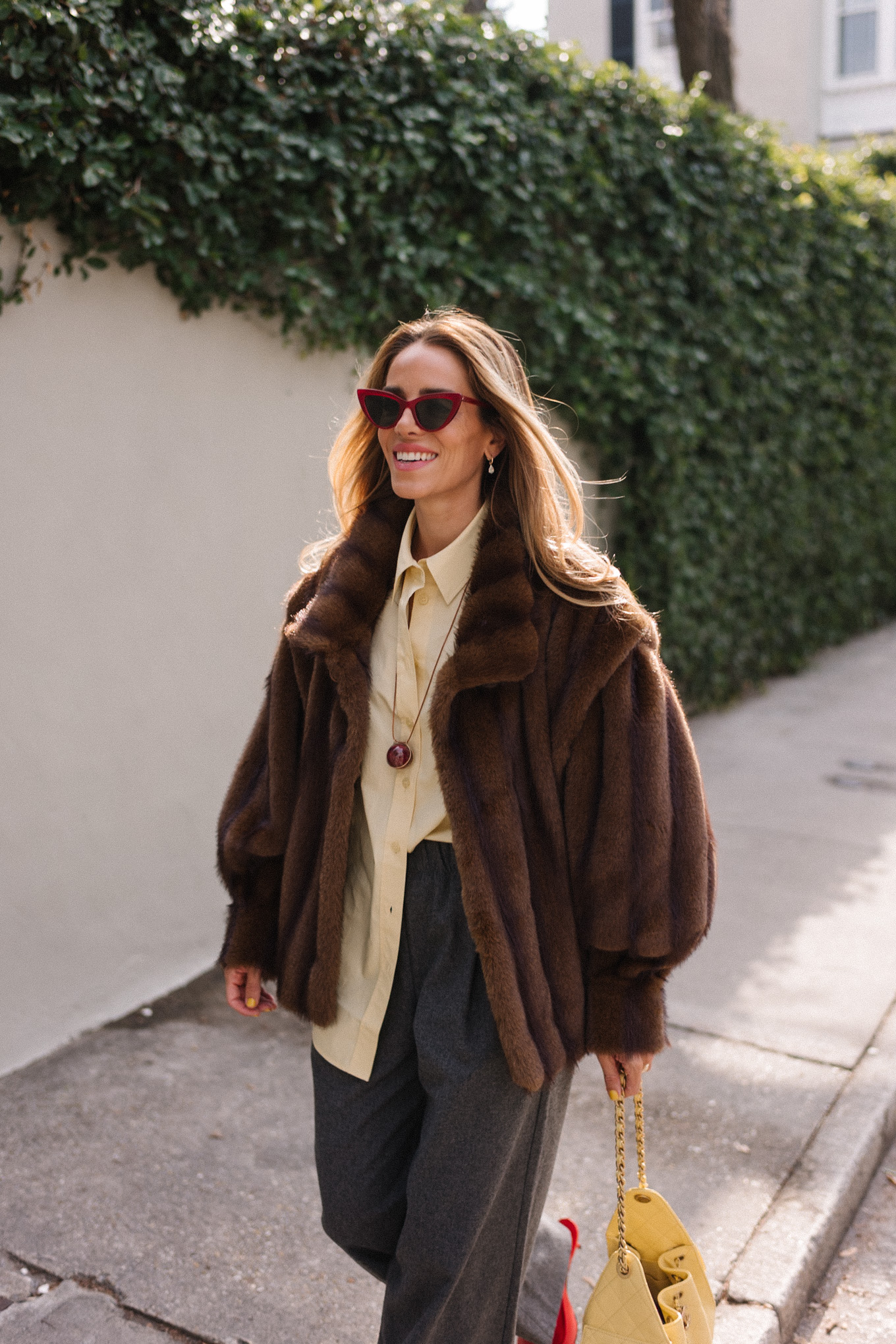 Manteau en fausse fourrure marron, chemise boutonnée jaune, pantalon gris, sac fourre-tout jaune, ballerines rouges