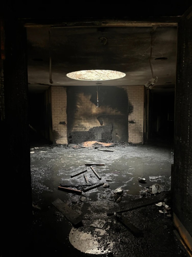 A dark, charred room covered in ashes and debris with smoke marks on the brick wall, ceiling and floor. A circular hole in the ceiling brings warm light into the center of the room.