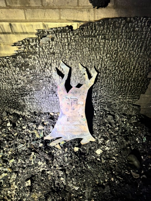 A metal sculpture in the shape of a tree with the Hebrew word for “alive” lies against a wall surrounded by charred material.