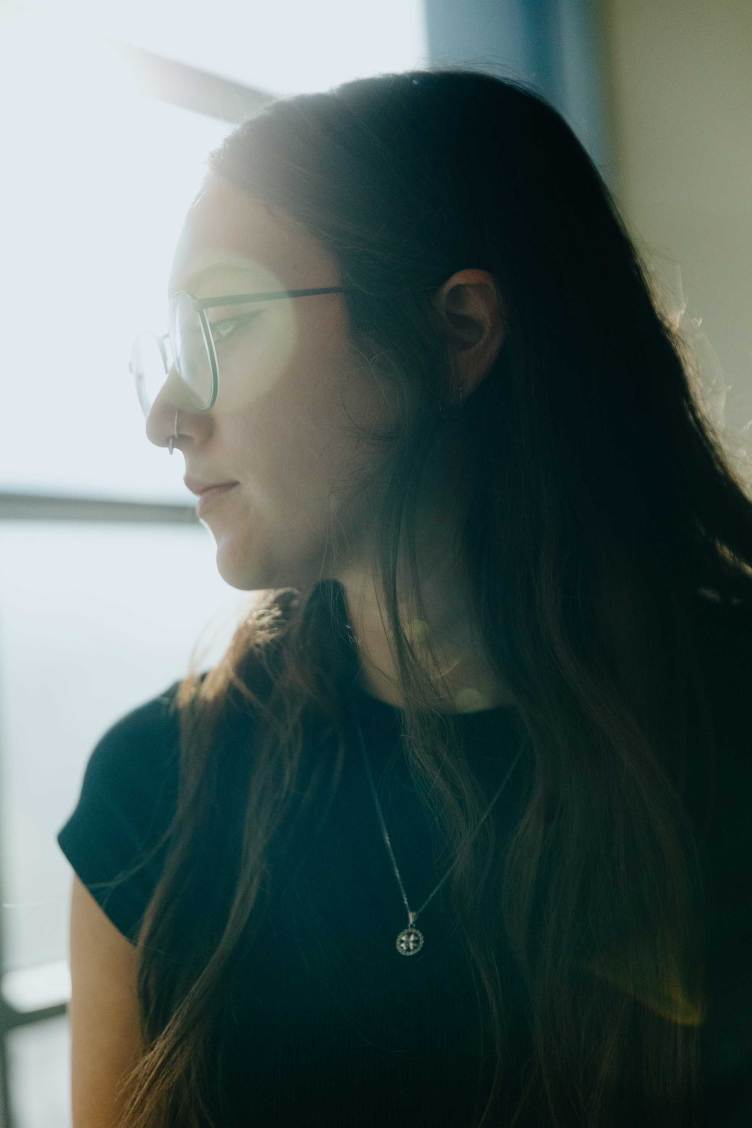 A portrait of a woman with long brown hair and round eyeglasses, wearing a black T-shirt.