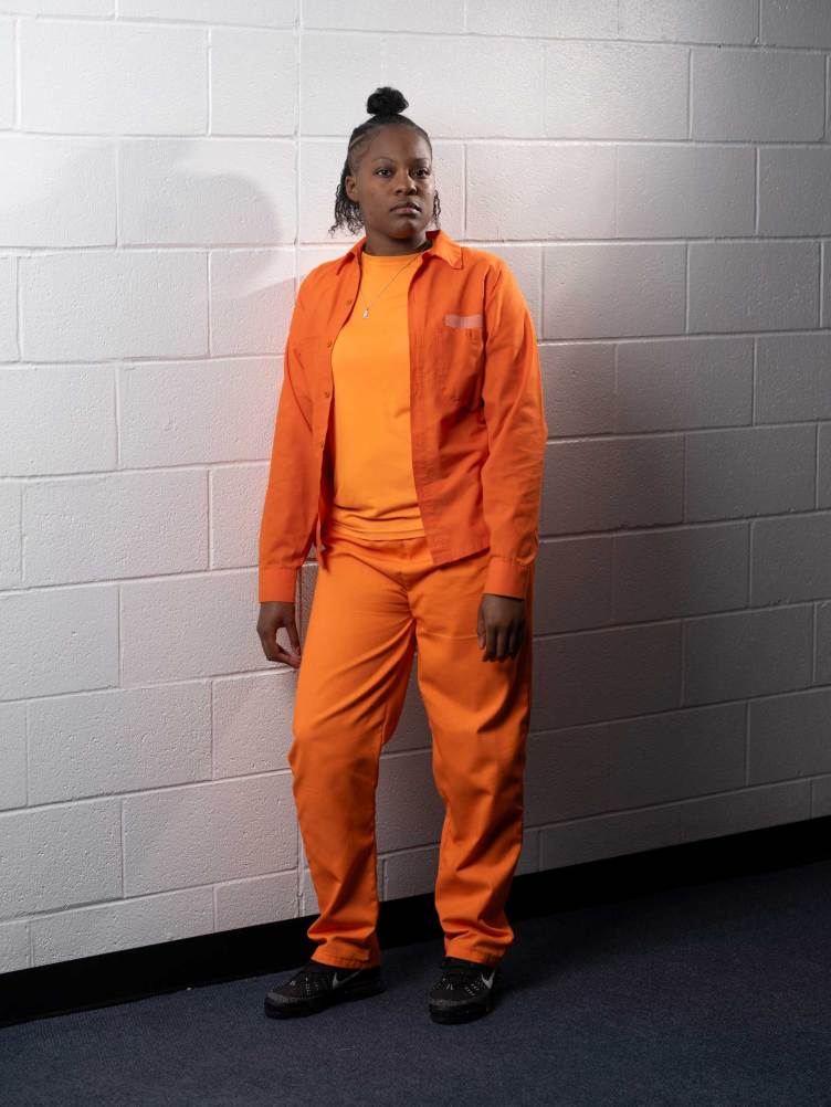 A woman with her hair in a top knot wearing an all-orange outfit stands against a white, painted cinder block wall.
