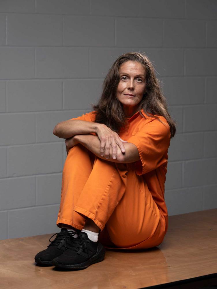 A woman with long brown hair sits on a wooden table while wearing an all-orange outfit in front of a white, painted cinder block wall.