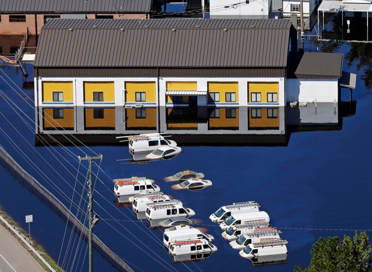 Vans and sedans sit half-submerged in dark blue water next to a building with yellow paint and windows reflected in the water.