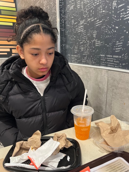 A young girl with her black hair pulled back, wearing a black jacket, looks listless while sitting at a McDonald’s with food wrappers and a half-empty orange drink on a table in front of her. 