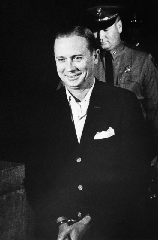 A black and white photo of a handcuffed man wearing a black suit and white shirt grins widely as a police officer stands behind him.
