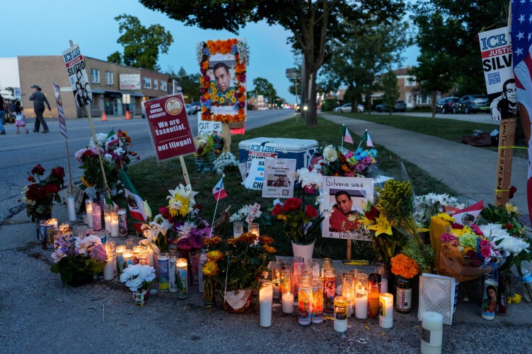 Lit candles and flowers lined up along the curb of a grassy area between the street and sidewalk.