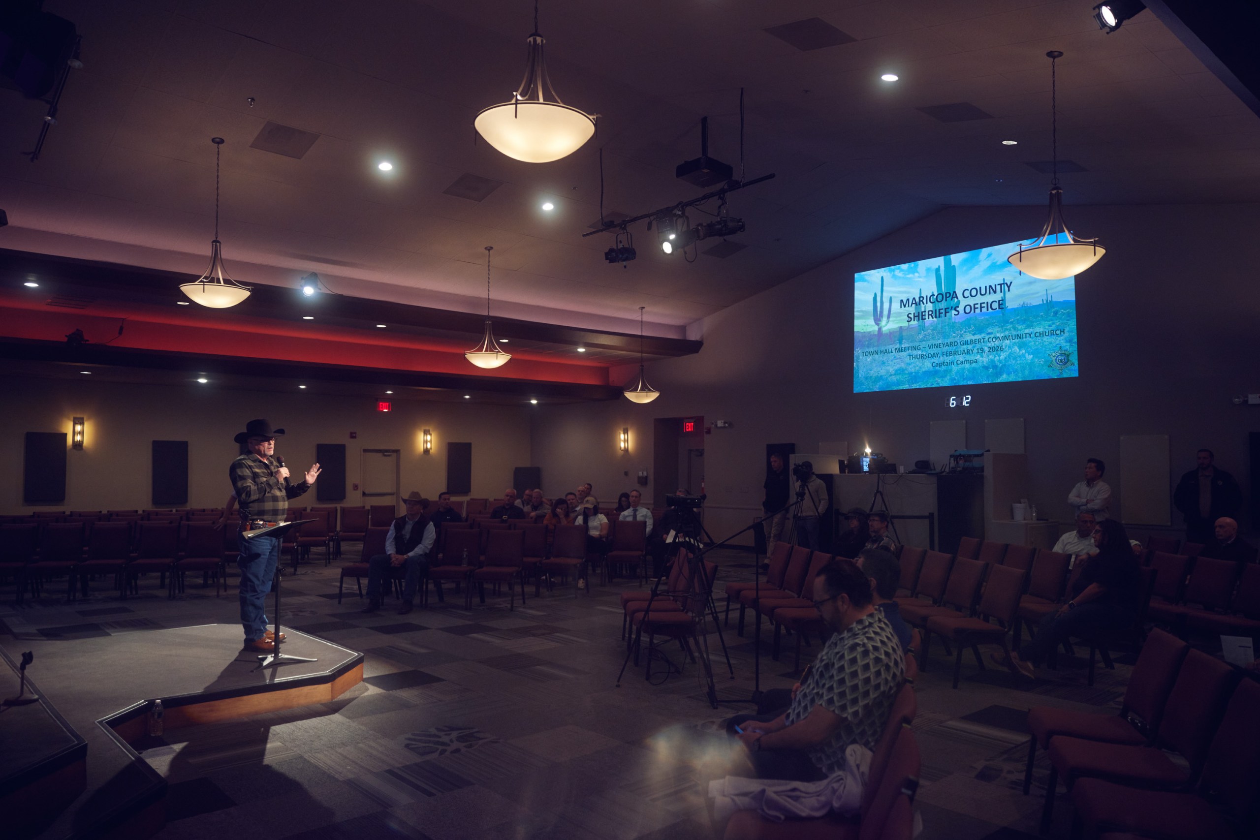 A man wearing a flannel shirt, jeans and a cowboy hat speaks into a microphone from a stage to a crowd of people in a large, dimly lit meeting space with a projector display on the back wall.