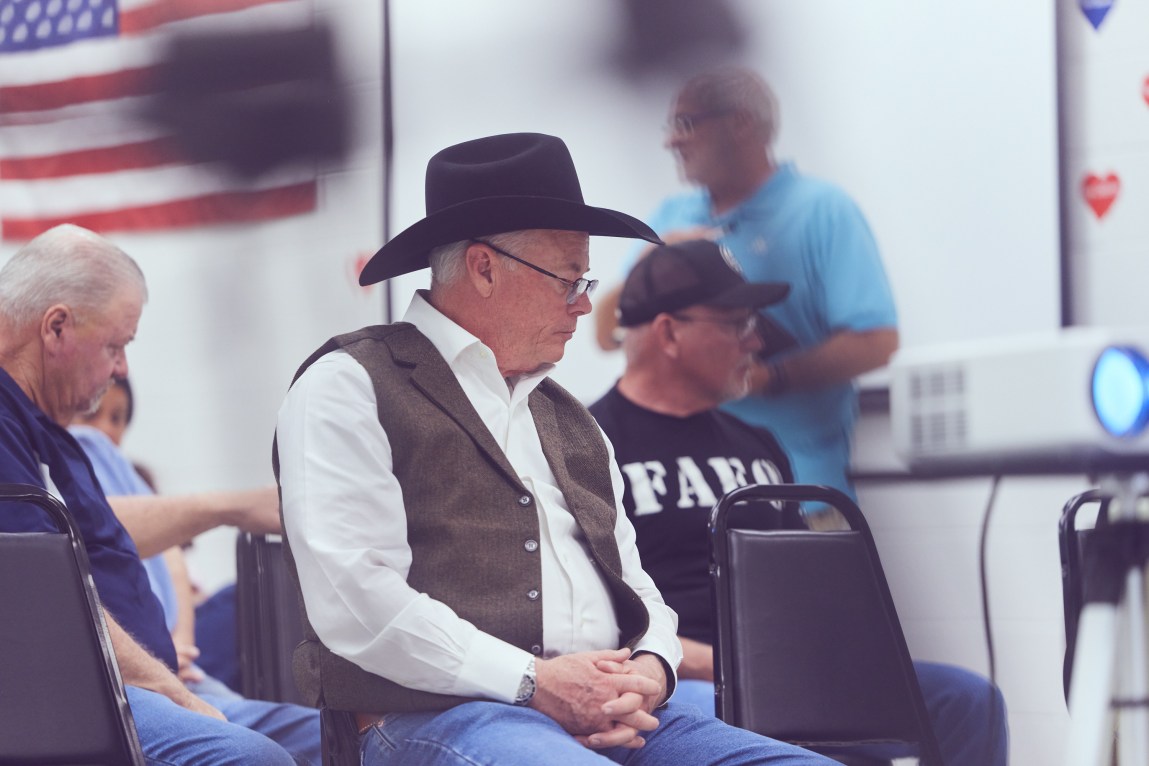 A seated man wears a black cowboy hat, tan vest, white dress shirt and blue jeans. Other people are seated behind him, and off to the side a projector glows.
