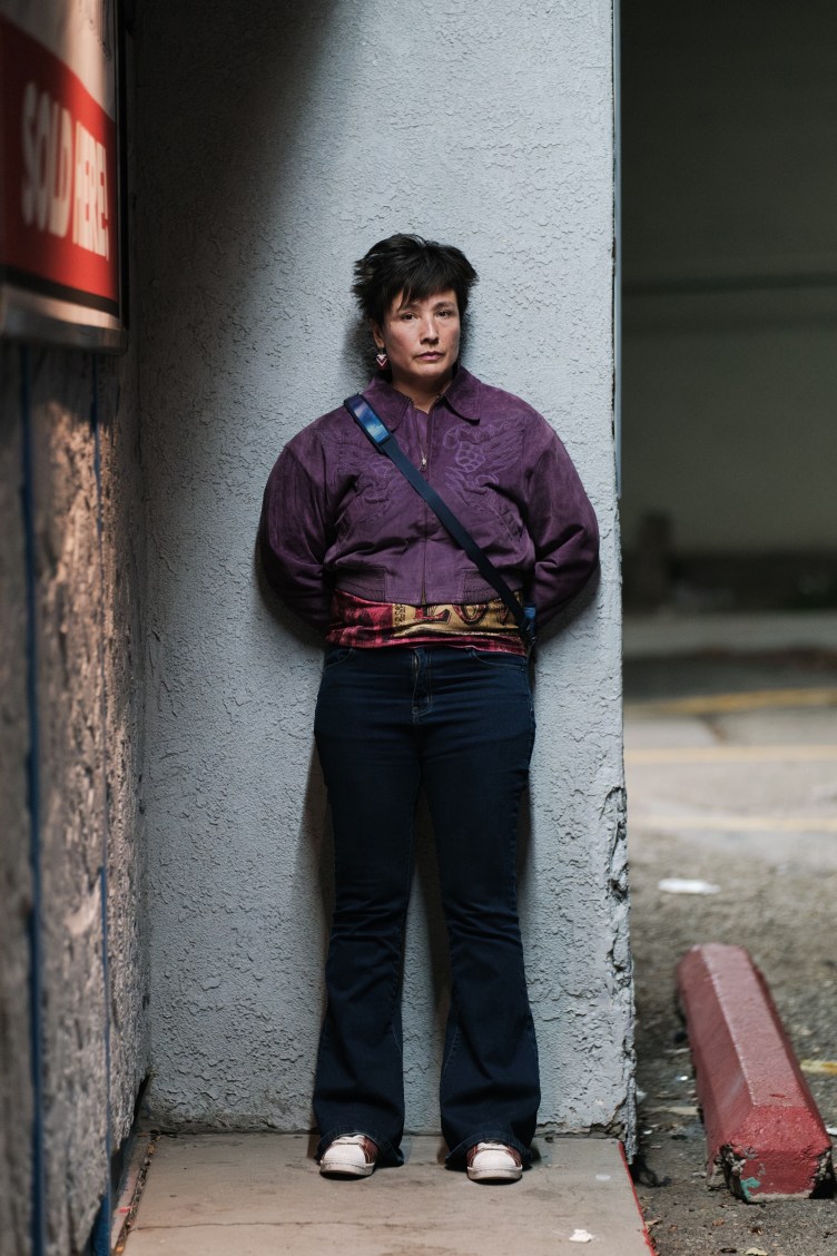A woman in a purple jacket and black pants stands against a blue wall at night.