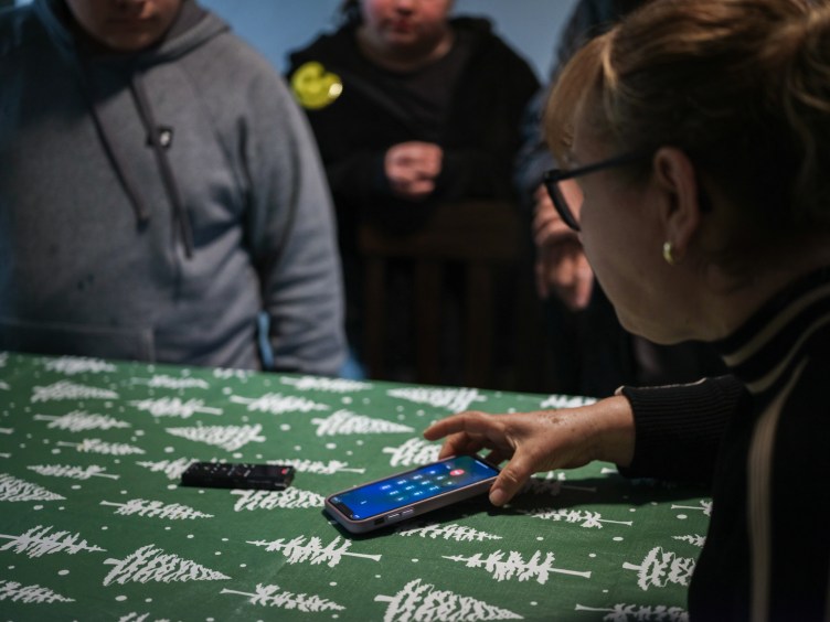 A woman holds a cellphone on a table, which has a green tablecloth with a white Christmas tree pattern, as three individuals stand in the background.