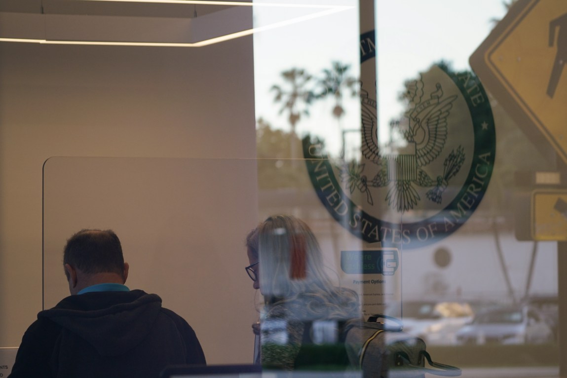 A man and woman are seen from behind in a building through a large window with the decal of U.S. federal seal.