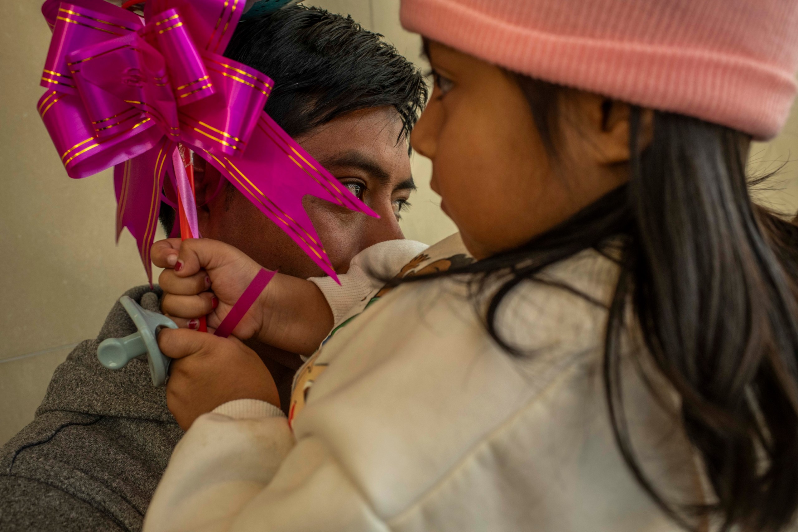 A man with black hair and a gray sweatshirt, with his face obscured, holds a young girl with long black hair, wearing a pink beanie and white sweatshirt and holding a pink ribbon and blue pacifier.