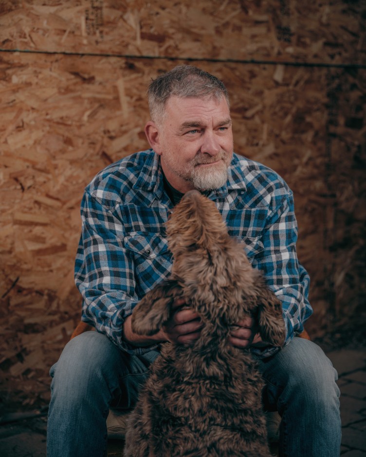 A seated man, wearing a blue checkered shirt and blue jeans, ruffles the fur of a dog’s neck.