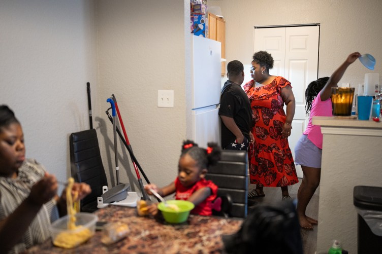 Two children eat pasta while sitting at a table. In the background a woman and another child look at the kitchen counter and another child reaches for a bowl.