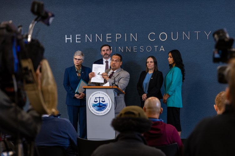 Un homme en costume brandit un morceau de papier alors qu'il se tient derrière un podium avec un petit microphone, flanqué de trois femmes et d'un homme, également en costume, avec des journalistes et des caméras assis devant eux.