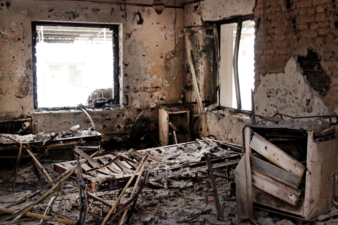 A room with two empty windows filled with rubble and ash. Plaster has been knocked off areas of the brick walls, two charred beds stand in the middle of the room and two bent and broken metal carts stand nearby. 