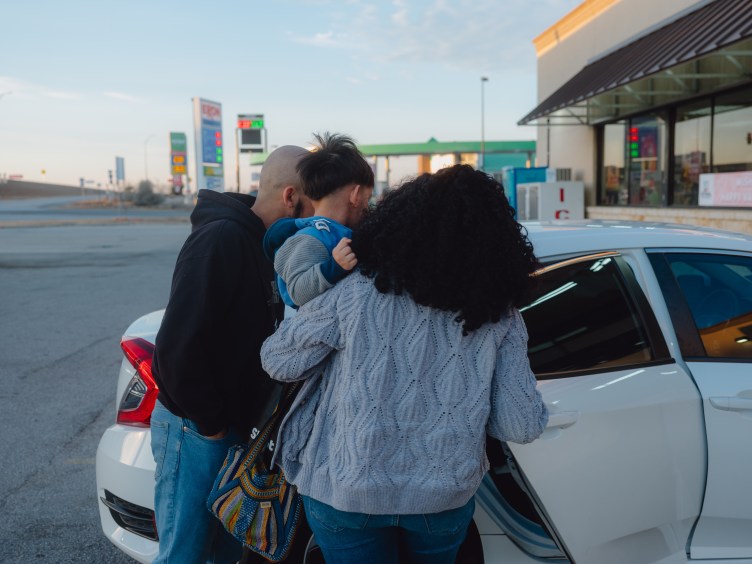 A man and a woman place a young boy in the back seat of a white car, outside a gas station.