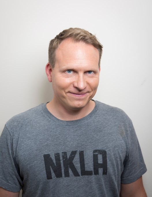 Un homme vêtu d'un T-shirt gris sourit à la caméra sur un fond blanc uni.