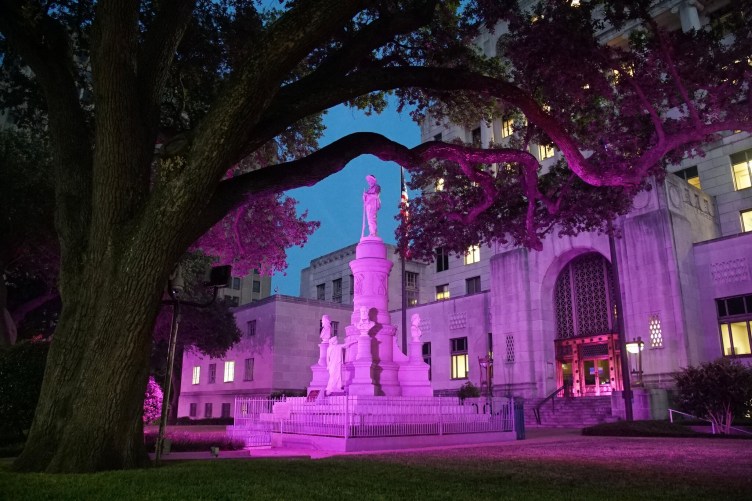Pink light illuminates a statue, which includes a soldier and the busts of several men, in front of a large government building.