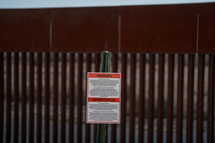 A sign on a post stating that the area is a military zone in English and Spanish, in front of the slotted steel bollard U.S. border wall with Mexico.