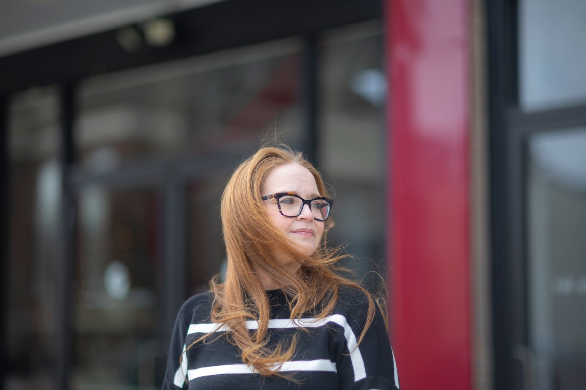 A woman with windswept hair and glasses, wearing a black-and-white striped top, stands in front of a blurred building and looks away from the camera.