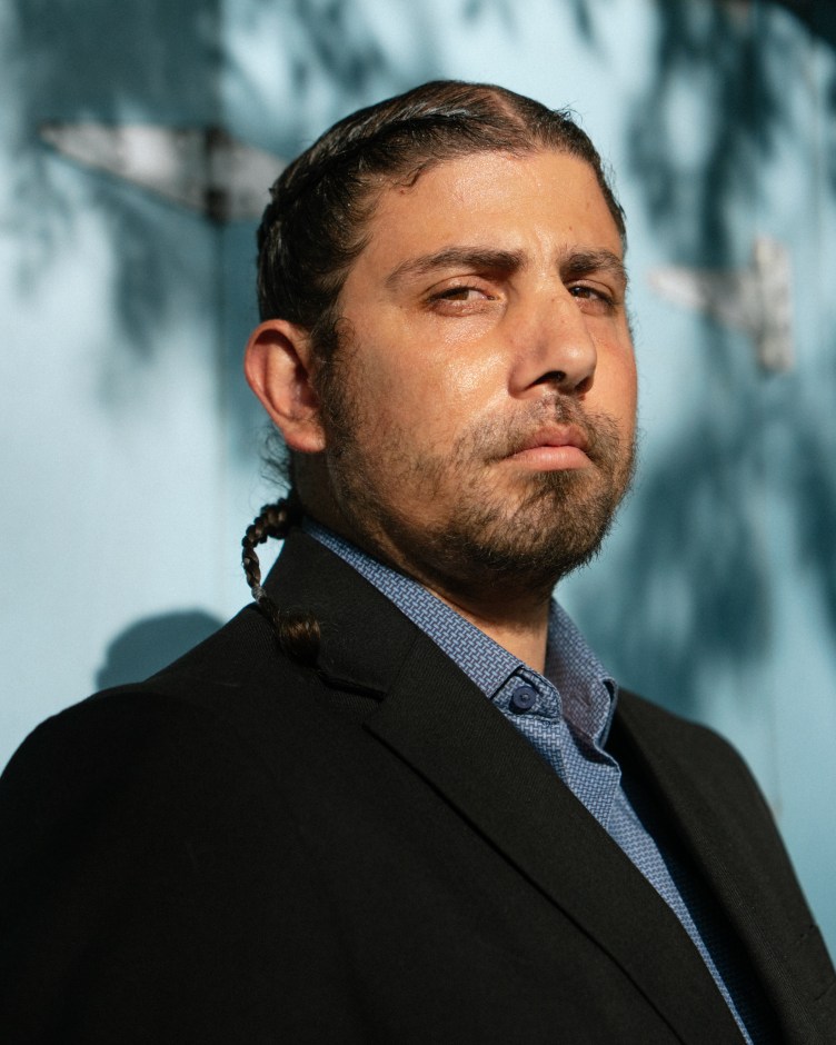 A man with his hair in two small braids looks into the camera. He’s wearing a collared shirt and suit jacket.