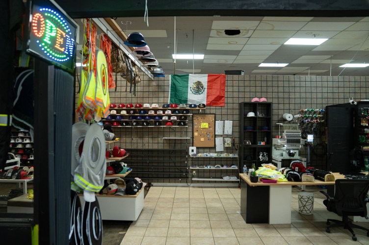 An empty store displays hats and other goods, with a prominent Mexican flag displayed and a colorful “Open” sign.