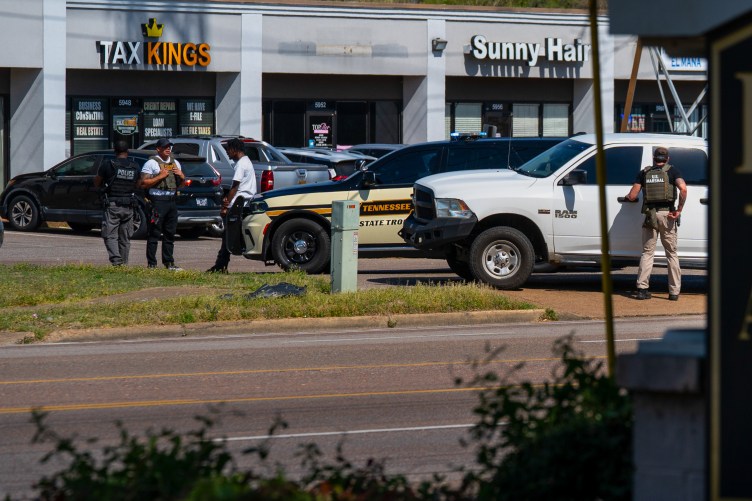 Men with vests reading “Police” and “US Marshal” conduct a traffic stop, speaking with a man outside of their cars in a strip mall parking lot.