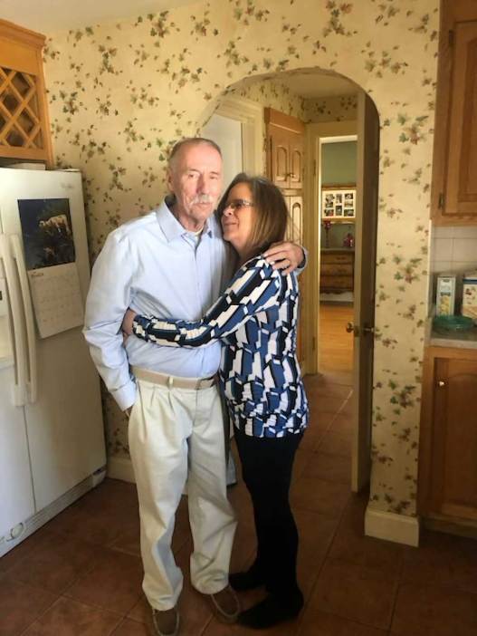 A man and a woman embrace in a hug, standing in a sunlit kitchen.


