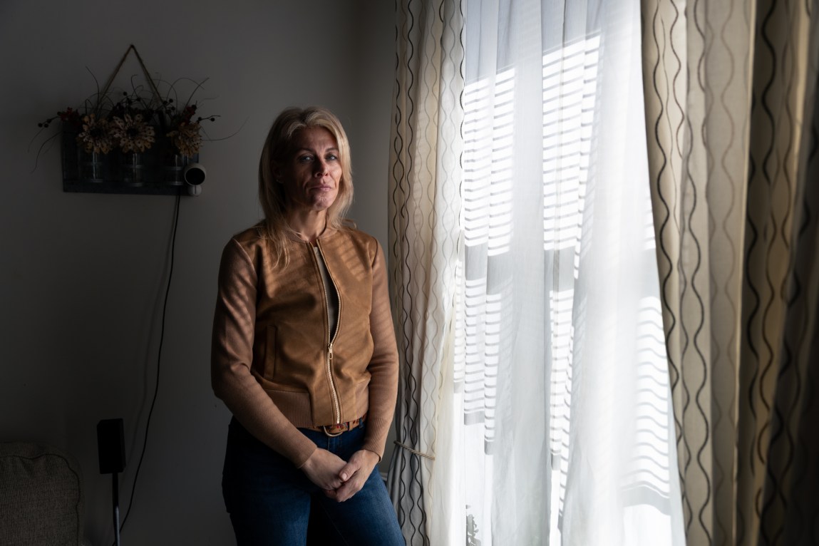 A blond woman wearing a brown jacket and jeans stands next to a window that has light streaming through the sheer white curtains. Hanging on the wall behind her is a basket of flowers.