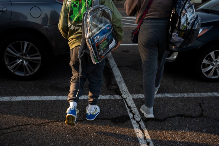 Two students holding backpacks walk away from the camera in a parking lot.