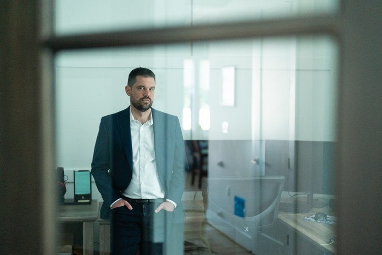 A man in a suit stands behind a glass pane. He is looking directly at the camera with his hands in his pockets.