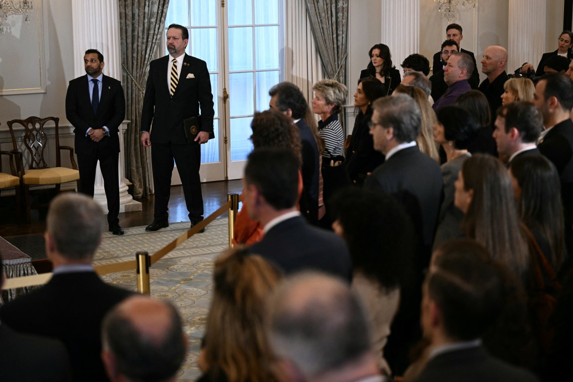 At the State Department, two men in suits stand behind a golden rope separating them from a crowd of people observing something off camera.