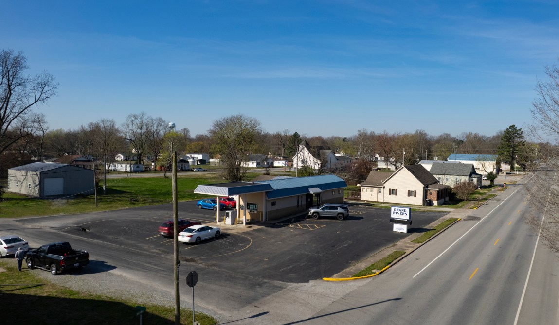 A small, drive-through bank building in a small-town setting, with roads and parked cars in the foreground and houses, other buildings and trees in the background.