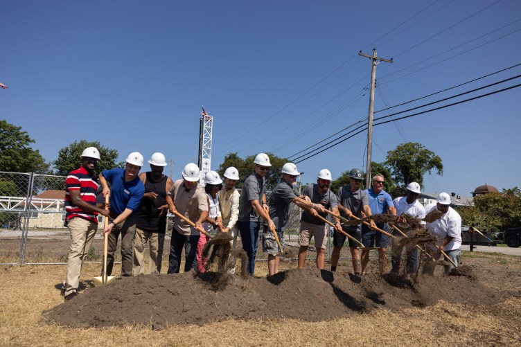 Treize personnes portant des casques de sécurité se tiennent en rang et pelletent un tas de terre à l'extérieur. Des lignes électriques et une structure qui ressemble à une tour, faisant partie d’une imprimante 3D géante, sont visibles en arrière-plan.