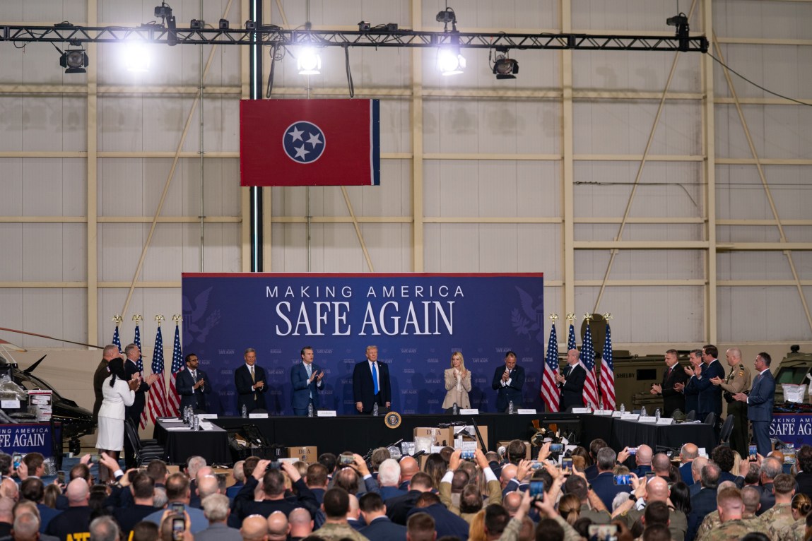 President Donald Trump stands in front of a large, cheering crowd, flanked by clapping members of his administration, in a hangar. Behind him is a banner that reads “making America safe again.”