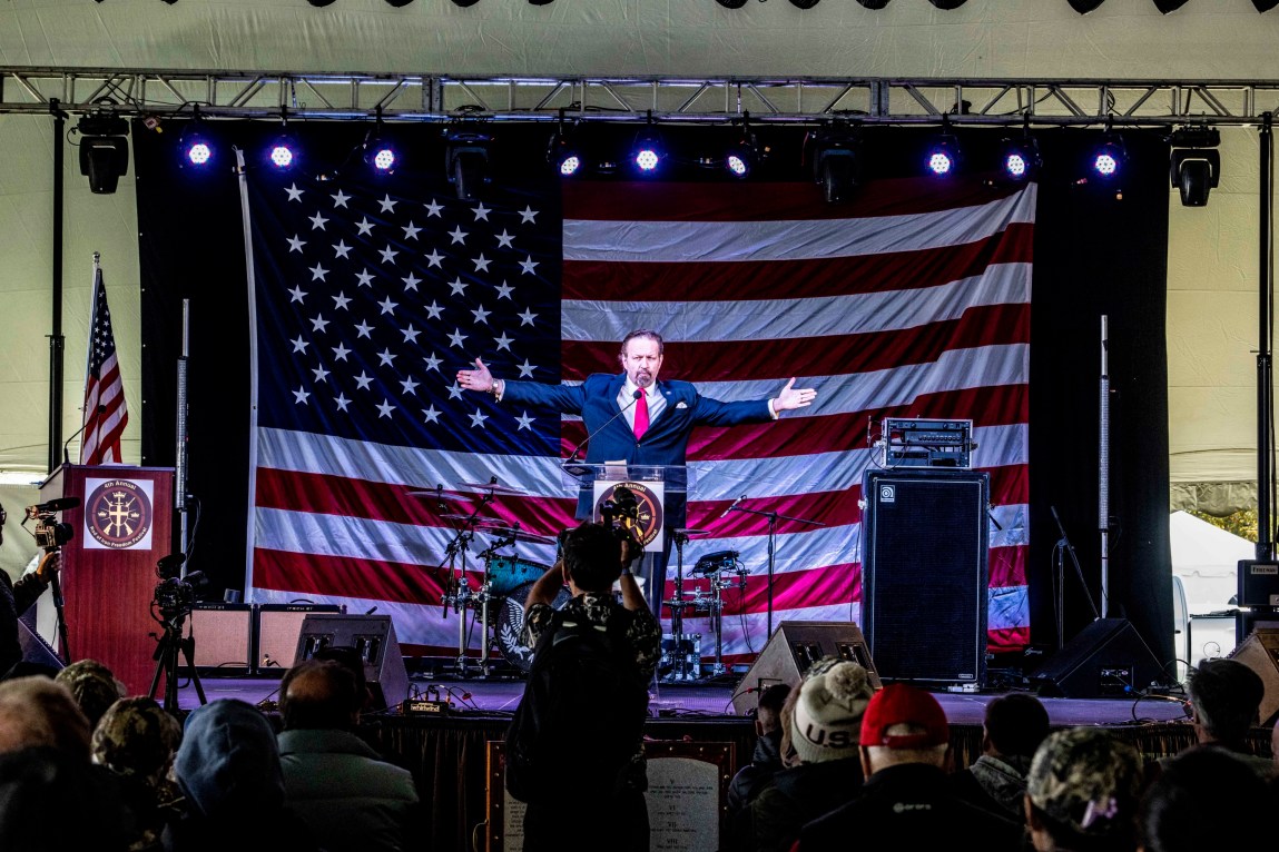 A man wearing a suit is speaking behind a lectern in front of a large American flag. His arms are open in a T shape as he addresses the crowd.