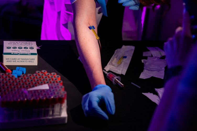 An arm with a butterfly needle stuck into it. The person’s gloved hand rests on a black table near a rack of vials, a box of medical tape and crumpled paper packaging from medical supplies.