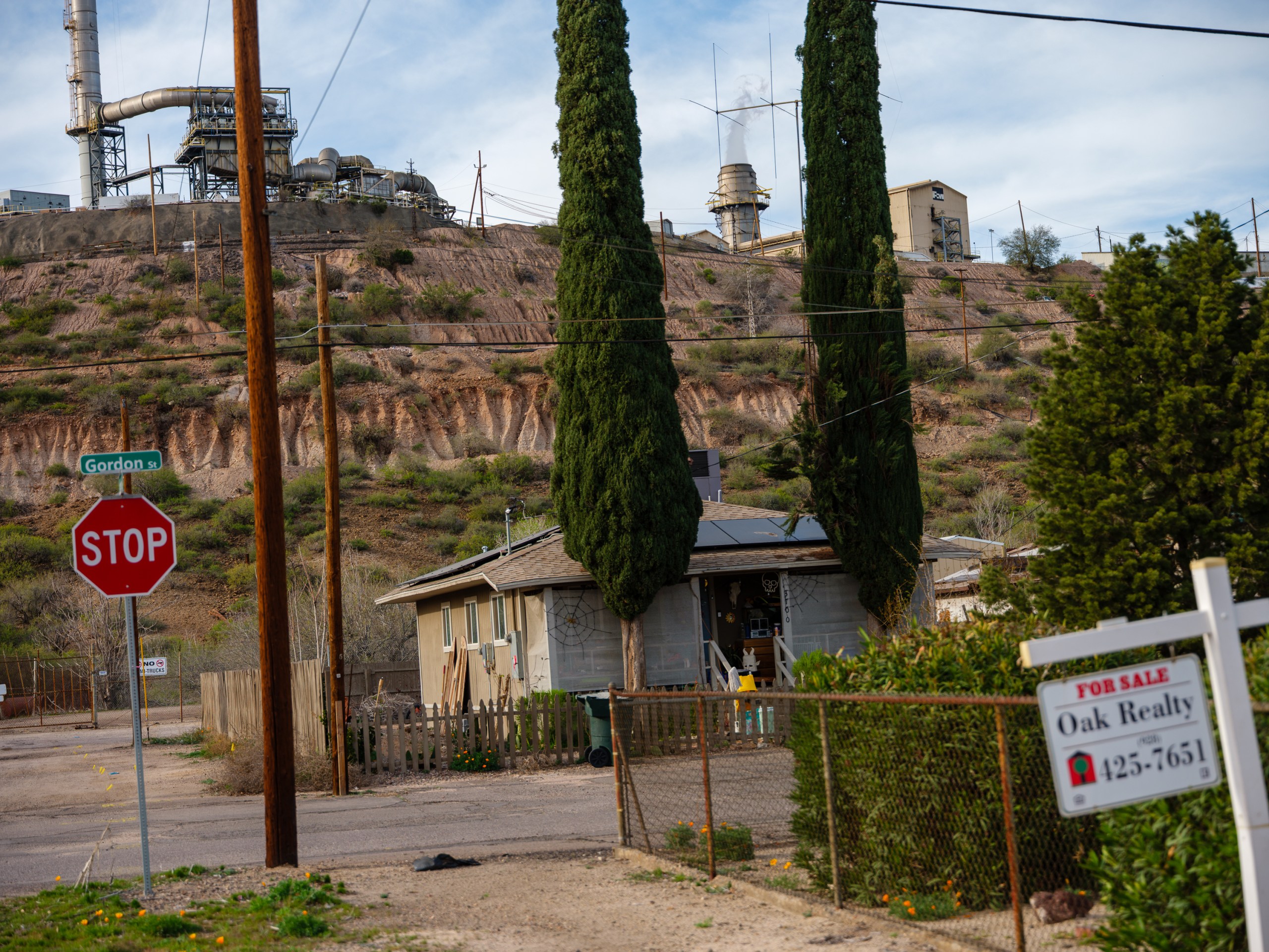 Smoke stacks rise from a smelter, sitting on a hill above a small gray house near a red stop sign and a white for-sale sign.