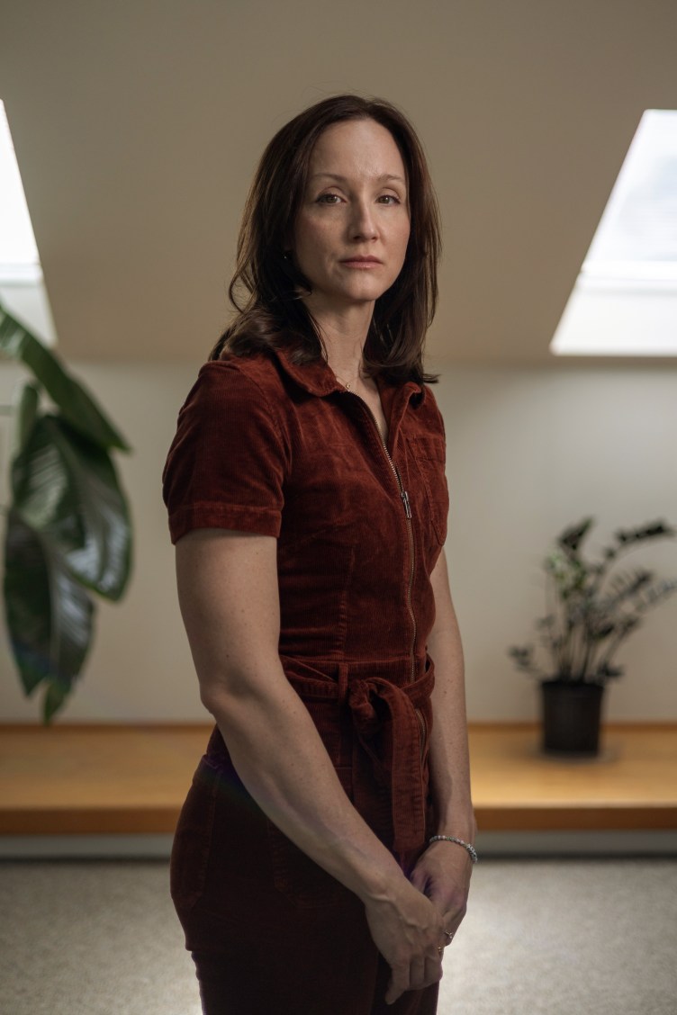 A three-quarter-length portrait of a woman with shoulder-length dark hair wearing a reddish-brown corduroy jumpsuit, standing indoors under angled skylights with houseplants in the soft-focus background.