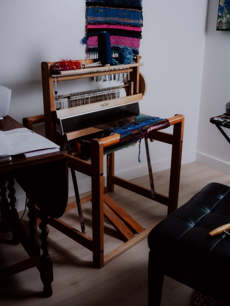 A large wooden loom, against a white wall in a room, with blue material in it.