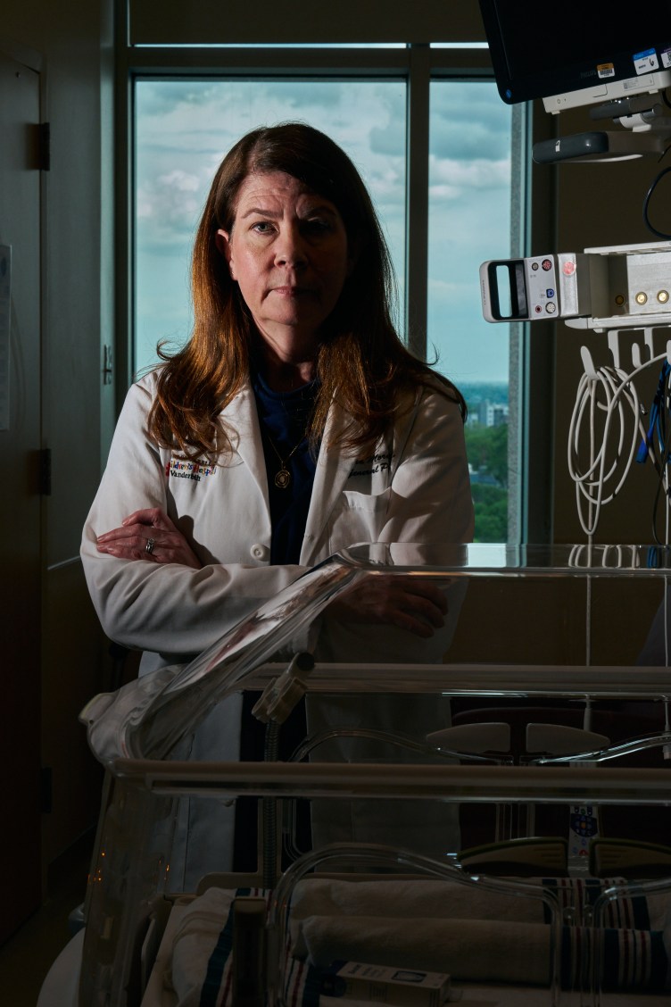 A woman with long red hair, wearing a white lab coat, stands with her arms crossed in a pediatric hospital room.